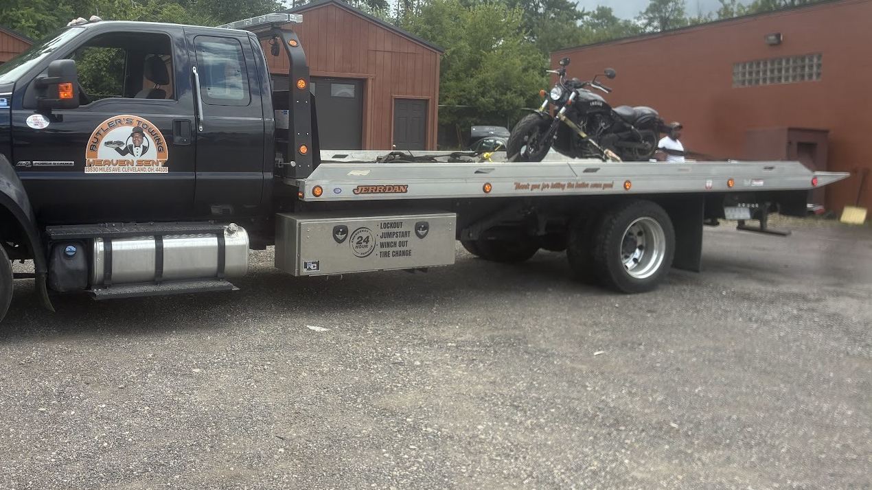 Butler's black flatbed tow truck with the Butler's Towing door badge, a motorcycle strapped on the bed, parked in a gravel lot.