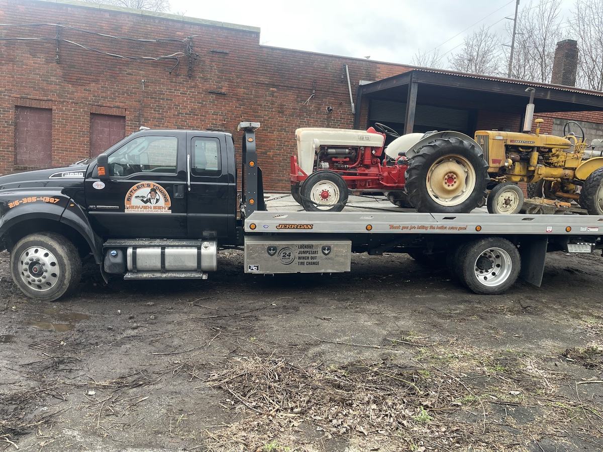 Butler's black flatbed carrying two old tractors, one red and cream and one yellow, beside a brick warehouse.